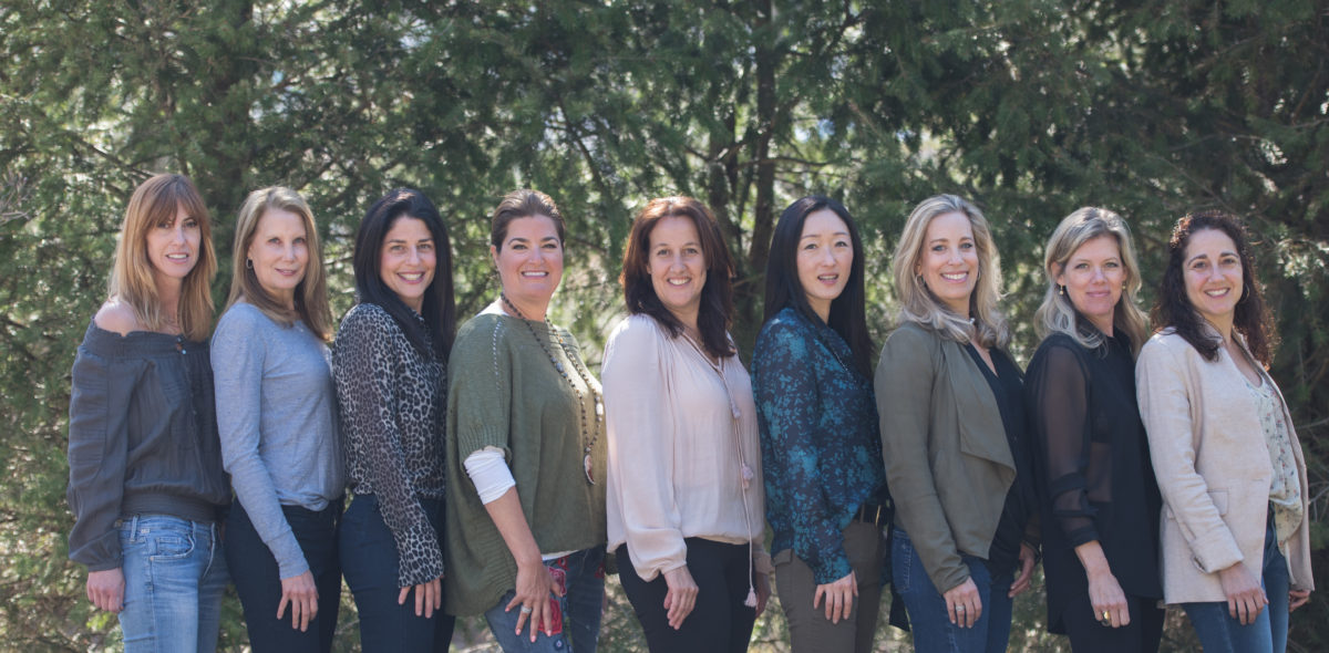 Six women standing side by side outdoors, smiling at the camera.